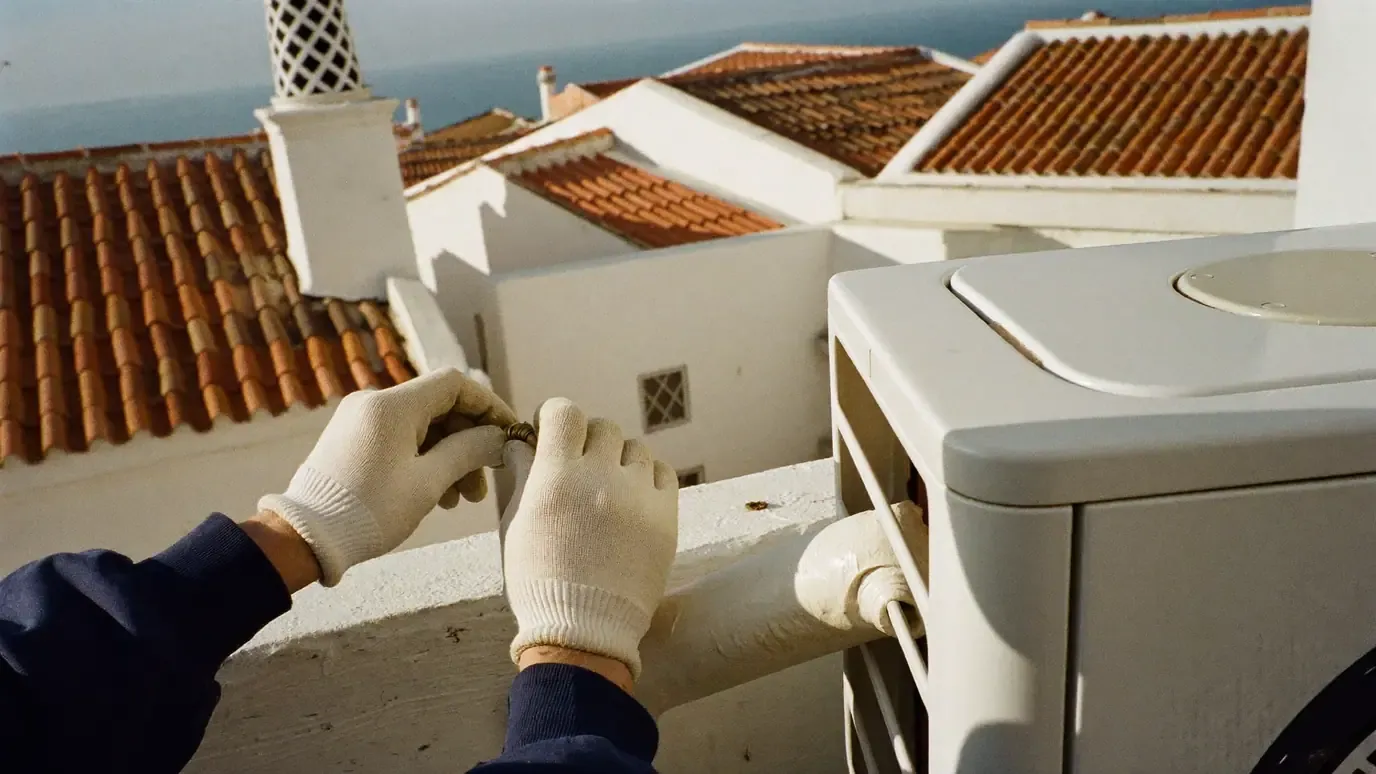 A technician's gloved hands thread a brass nut beside an air conditioning outdoor unit on a whitewashed rooftop terrace, with an insulated pipe running along the parapet, terracotta roofs, a lattice chimney and the sea behind