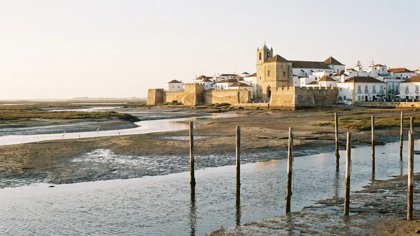 Faro old town with its stone walls and cathedral tower seen across the Ria Formosa at low tide, mooring posts in the foreground