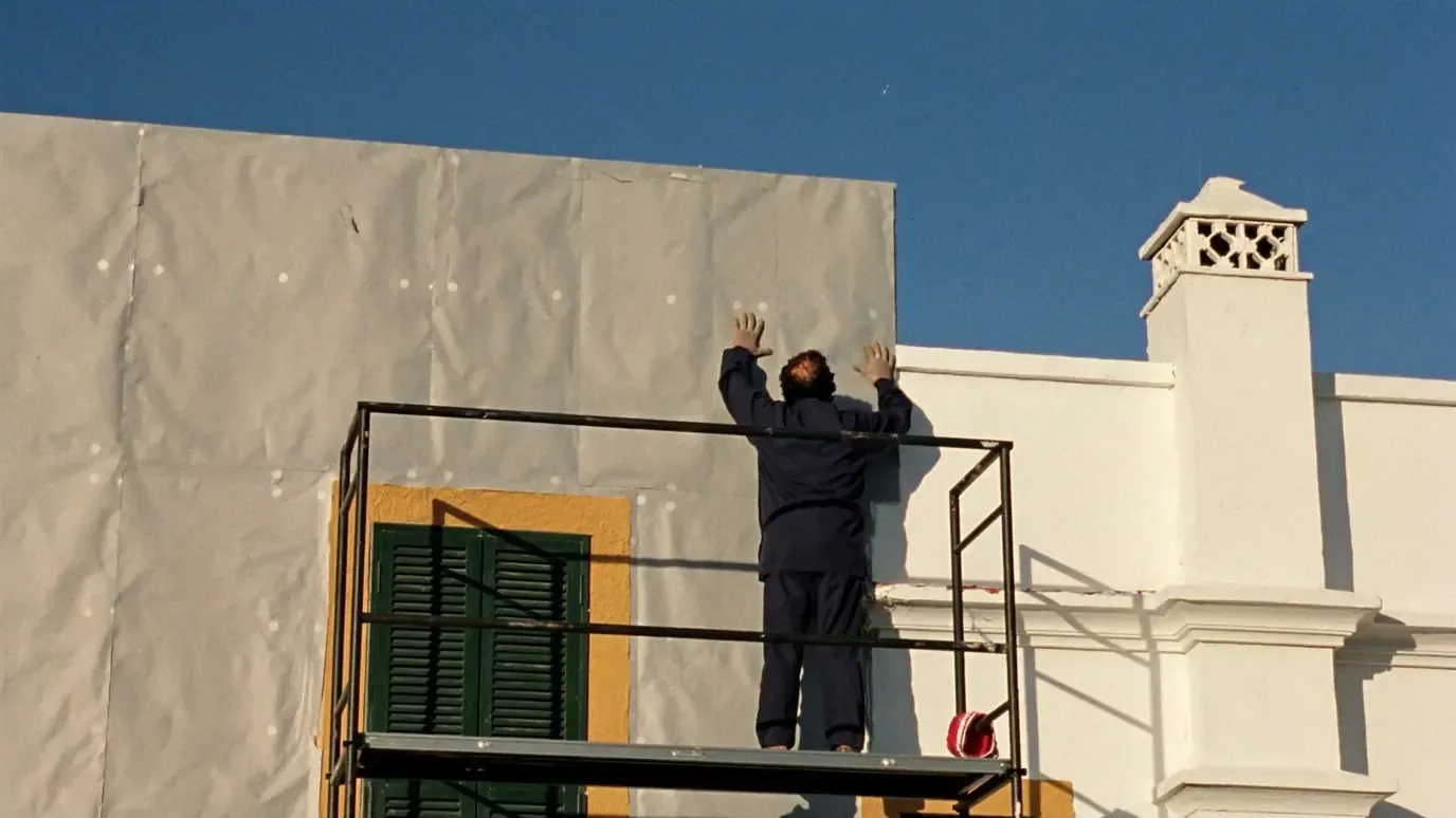 An installer seen from behind on a scaffold presses a grey insulation board fixed with white anchor discs onto the facade of a whitewashed house, beside a window with an ochre surround and green shutters