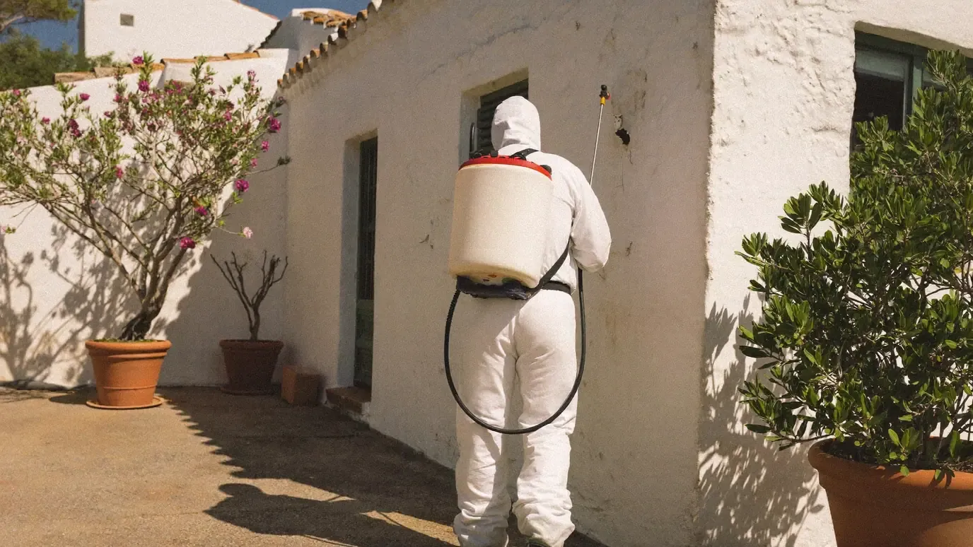 Pest control technician in a coverall using a backpack sprayer along an exterior wall
